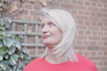 A woman with blonde hair wearing a red jumper is shown outside in front of a brick wall. She looks up and to her right.